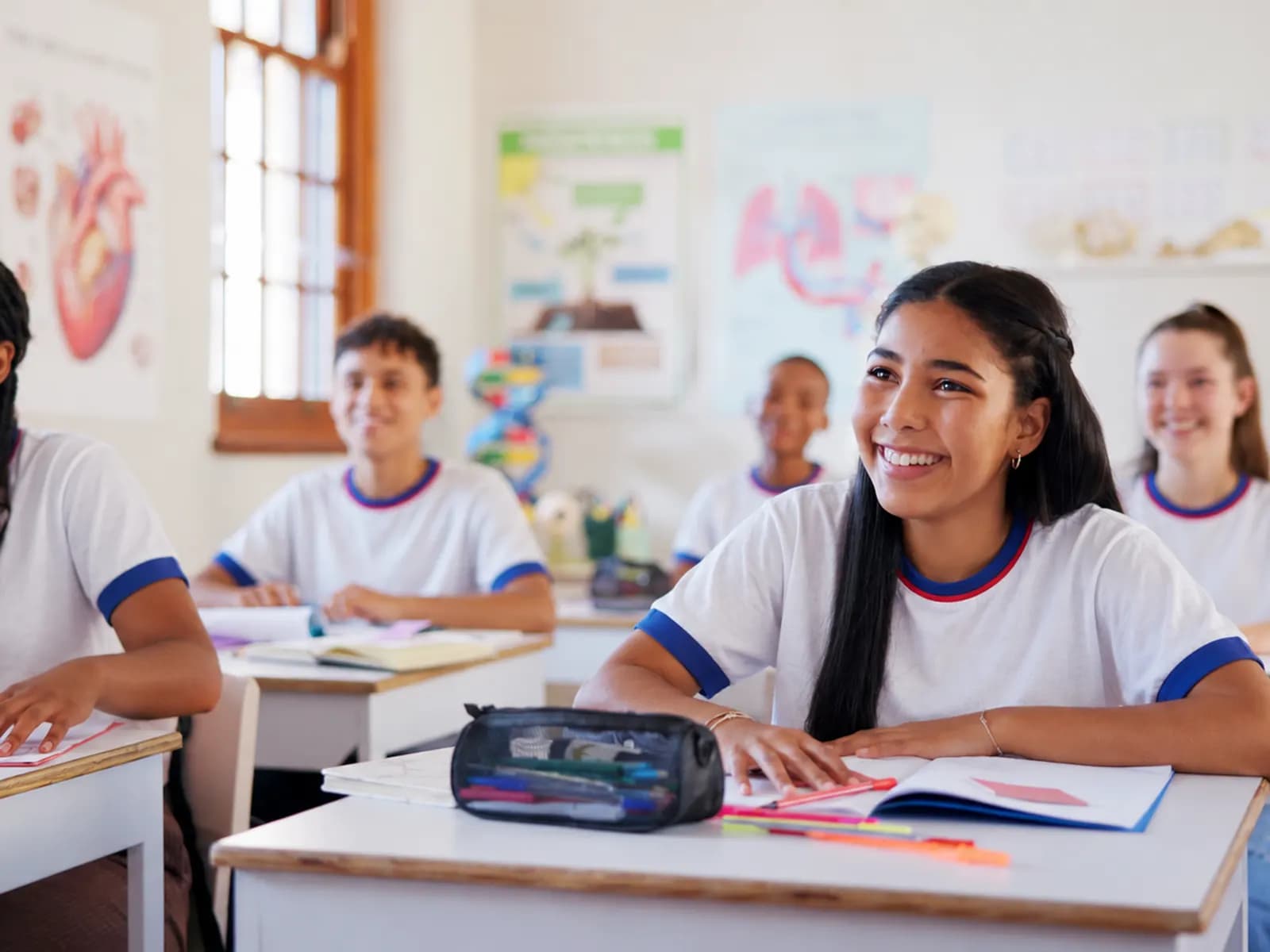 Estudantes sorrindo em sala de aula, com livros e materiais sobre a carteira