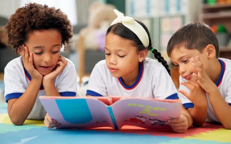Três crianças de uniforme lendo um livro ilustrado juntas